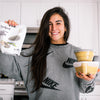 Lauren in a bright kitchen holding a bag of chia seeds and prepared meal containers, smiling while showcasing healthy meal-prep ingredients and nutritious food choices.