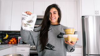 Lauren in a bright kitchen holding a bag of chia seeds and prepared meal containers, smiling while showcasing healthy meal-prep ingredients and nutritious food choices.