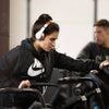 Lauren Fisher wearing headphones while using a cardio machine at the gym, focused on her workout with other gym members blurred in the background.