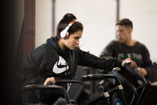 Lauren Fisher wearing headphones while using a cardio machine at the gym, focused on her workout with other gym members blurred in the background.