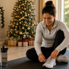 Woman tying her sneakers on a workout mat in a cozy living room decorated for the holidays with a Christmas tree, fireplace stockings, and warm natural light.